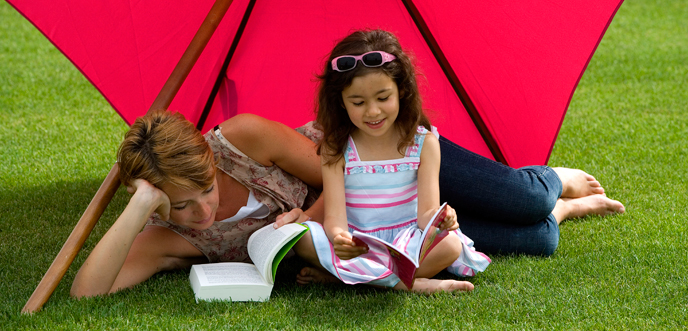 Fillette et sa mère faisnat de la lecture sur la pelouse à l'ombre d'un parasol rouge